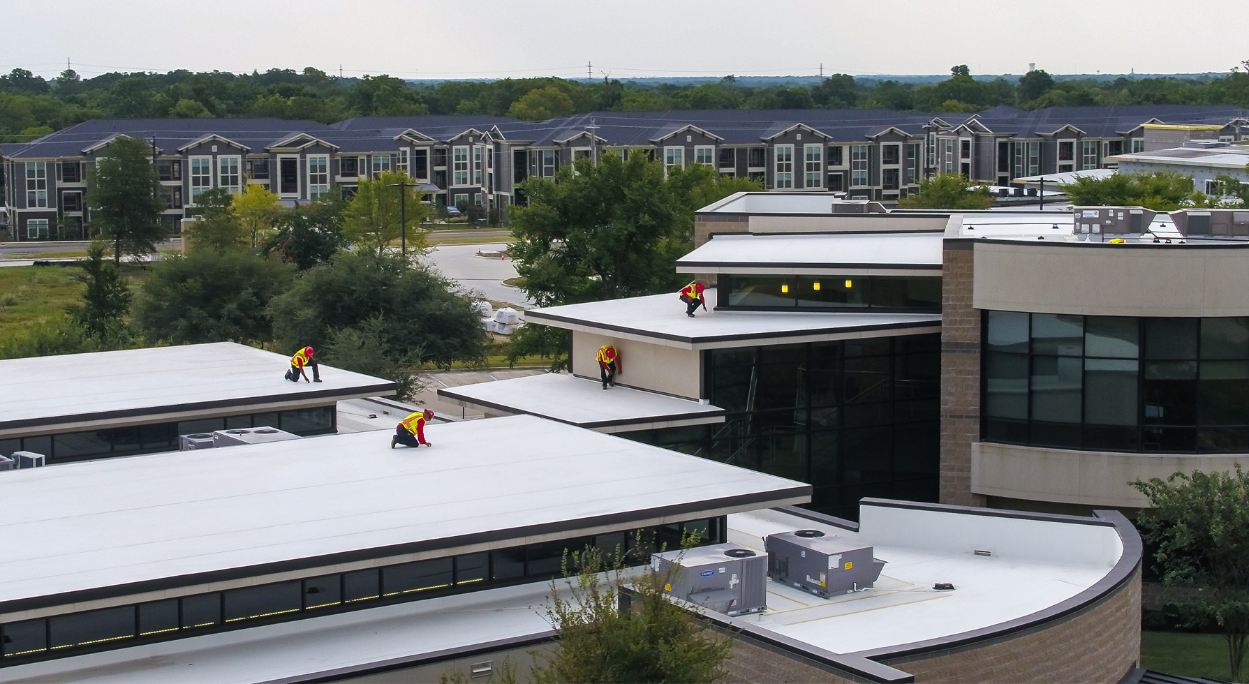 Elevated aerial view of five Schulte Roofing roofers, wearing red safety hardhats, florescent safety vests, signature Schulte Roofing red longs sleeve tops, jeans and work boots. They are kneeling or hunching over as they inspect a white six-tier commercial flat floor.